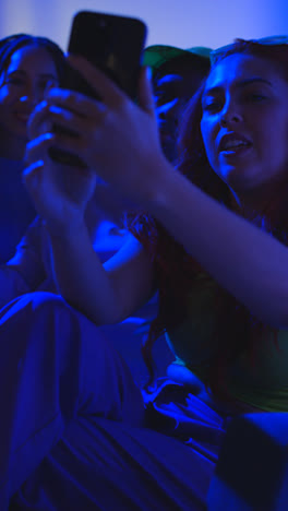 Foto-De-Estudio-De-Vídeo-Vertical-De-Un-Grupo-De-Amigos-De-La-Generación-Z-Sentados-En-Un-Sofá-Posando-Para-Un-Selfie-En-Un-Teléfono-Móvil-Por-La-Noche-Con-Luz-Intermitente